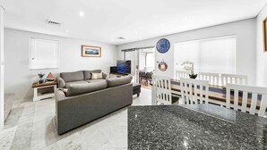 Ein modernes Wohnzimmer mit grauem Sofa, Fernseher und Essbereich mit weißen Stühlen und einem Holztisch.