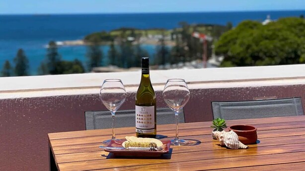 Ein Tisch mit einer Weinflasche, zwei Gläsern, einem Käseplatte und einer kleinen Pflanze auf einem Balkon mit Blick aufs Meer.