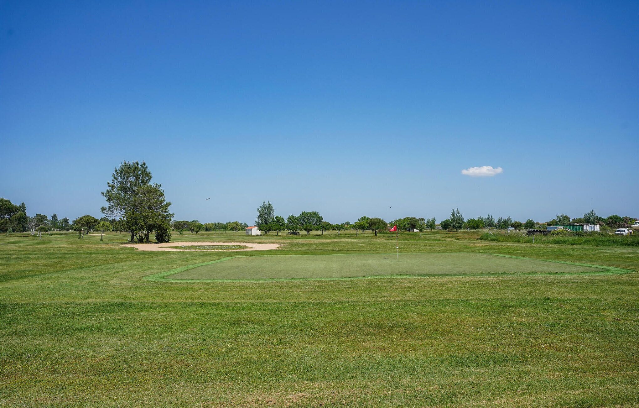 Golfkenttä, jonka yllä on kirkas taivas, ja jonka ympärillä on puita sekä kauempana pieni rakennus.