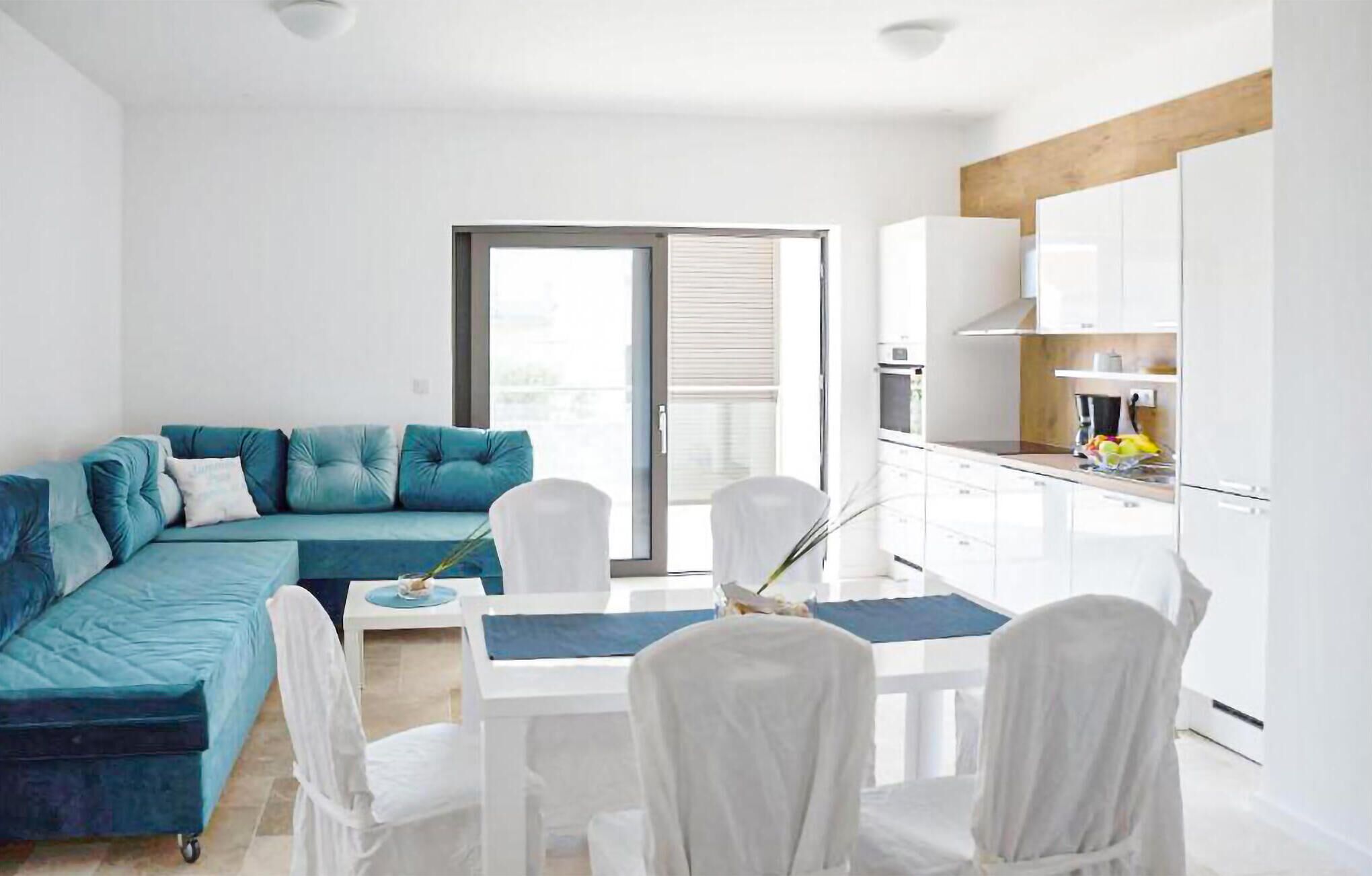 A modern apartment with a teal sofa, white dining chairs, a glass dining table, and a kitchen with white cabinets.