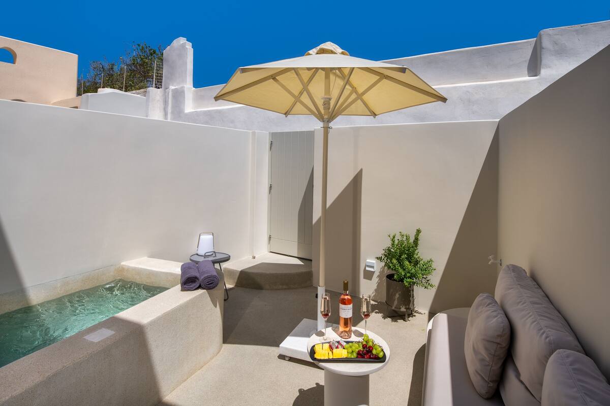 Un espacio al aire libre iluminado por el sol con una piscina, una mesa con una botella de vino, copas y fruta, y una sombrilla beige que da sombra.