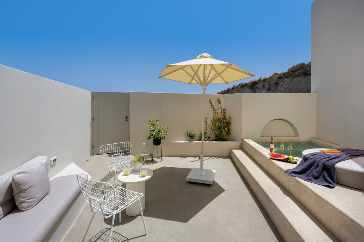 Un espacio exterior moderno con un sofá blanco, una silla de metal, una mesa con bebidas, una sombrilla, una pequeña piscina y una planta en maceta.