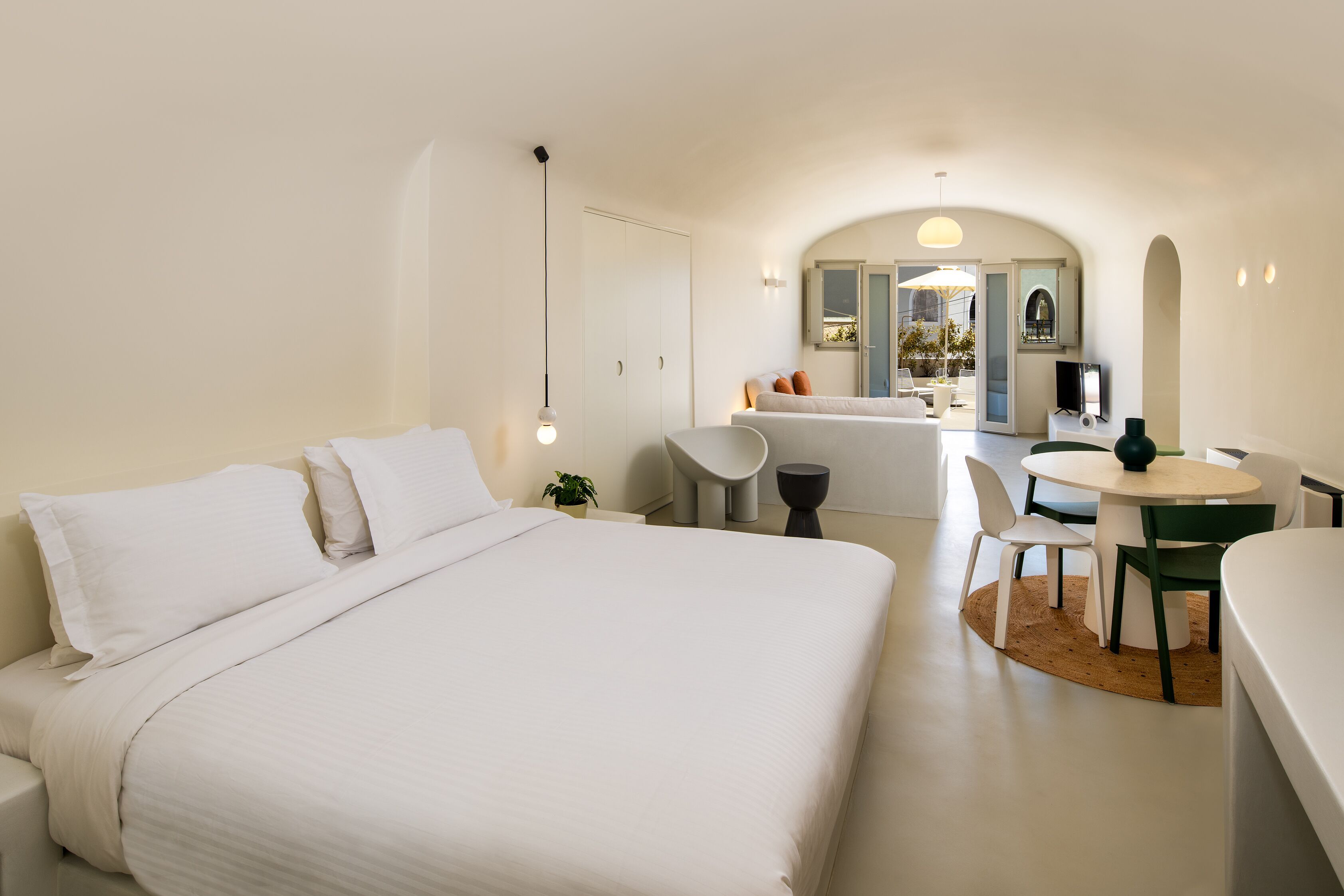 A modern hotel room with a large bed, a dining area with a round table and chairs, and a view of the outside through an arched window.