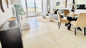 Een moderne woonkamer met een bank, eettafel, stoelen, salontafel, lamp, plant en een groot raam met uitzicht.