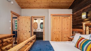 Un dormitorio de estilo cabaña con cama, cabecera de madera y vista a un baño con lavamanos y espejo.