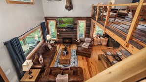 Interior acogedor de una cabaña con chimenea, sofás de cuero, muebles de madera y un ventanal con vistas a un bosque.
