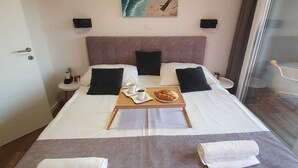 A neatly made bed with a tray of breakfast items, a painting of a beach scene, and a view of the sea through a window.