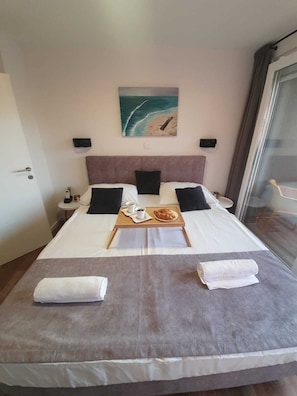 A neatly made bed with a tray of breakfast items, a painting of a beach scene, and a view of the sea through a window.