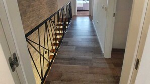 A hallway with a wooden floor, a black metal railing, and a view into a room with a balcony.