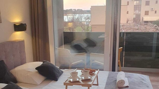 A hotel room with a bed, a small table with breakfast items, a towel, and a view of buildings through a glass door.