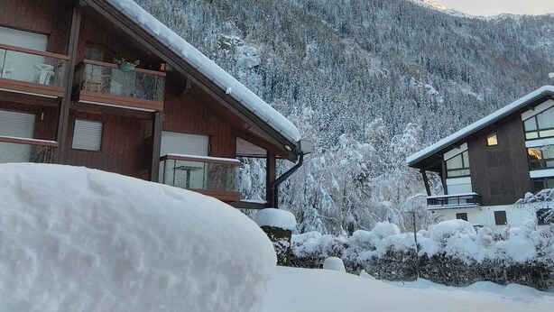 Una cabana de fusta amb grans finestres i un balcó, envoltada per arbres coberts de neu i muntanyes al fons.