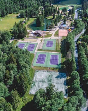 Eine Vogelperspektive auf einen Sportkomplex mit mehreren Tennisplätzen, umgeben von dichtem Wald und einem Fluss.