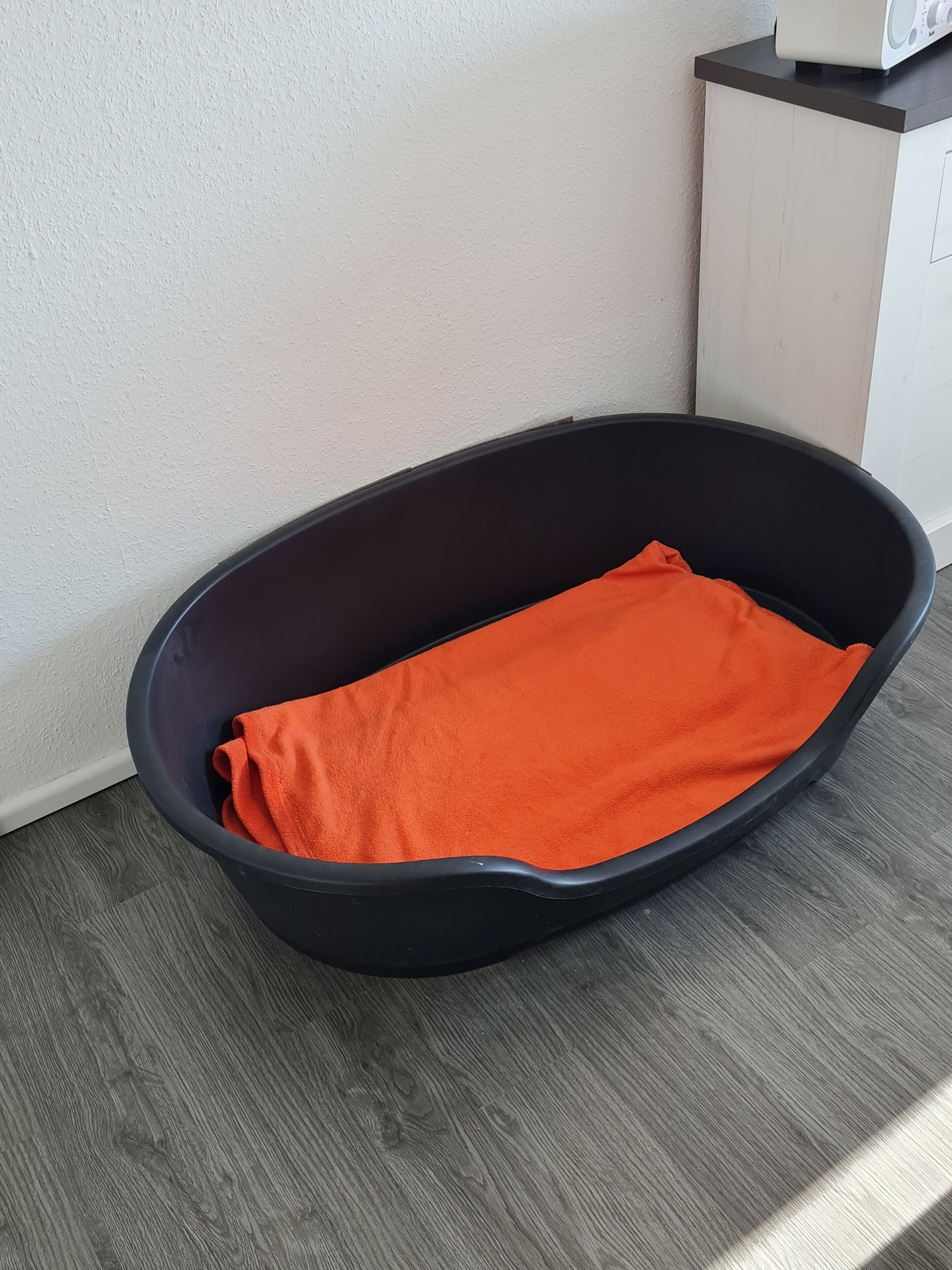 A black pet bed with an orange blanket on a wooden floor.