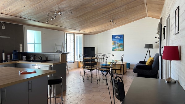 A modern kitchen with a wooden ceiling, a dining area with a TV, and a living space with a sofa and a lamp.