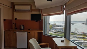 A compact kitchenette with a refrigerator, a sink, and a small countertop. There is a chair and a table near a window with a view of a busy waterfront with buses and a bridge. - Panorama Suite (Istanbul)