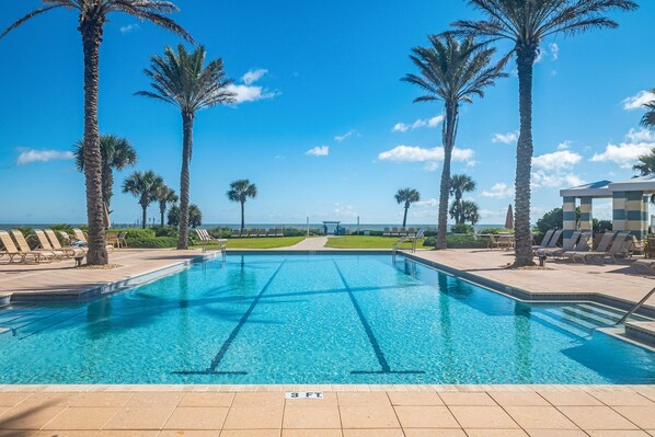 A swimming pool with clear blue water, surrounded by palm trees and lounge chairs, with a view of the ocean in the background.