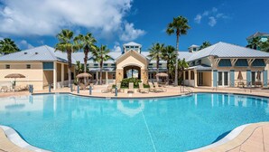 A large pool with lounge chairs and palm trees, surrounded by buildings with a Mediterranean architectural style.