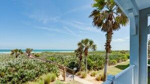 A coastal view from a balcony with a wooden deck leading to a sandy path, palm trees, and a beach in the background.