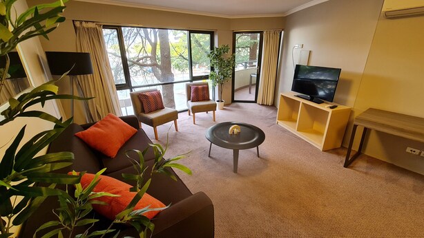 A modern living room with a sofa, armchairs, a coffee table, and a TV on a wooden stand. Large windows offer a view of trees and a balcony. - 1BR Executive Apartment, in City Centre (Braddon)