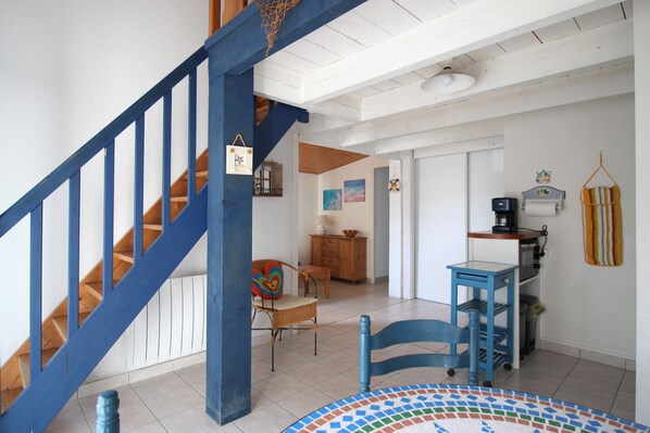 A staircase with blue railings, a wicker chair with a colorful cushion, a small table with a blue base, and a wall with a hanging basket and a framed picture.