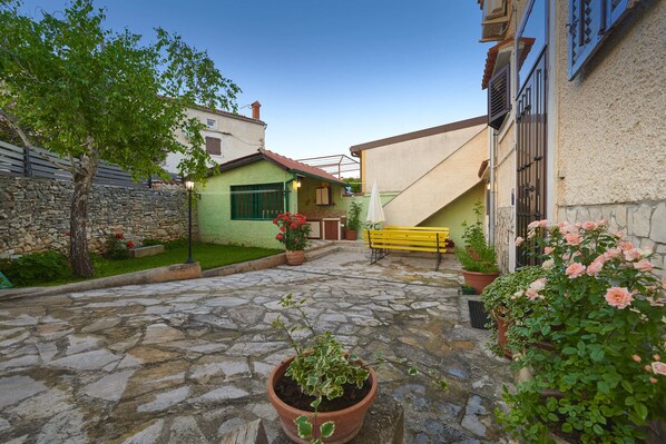 A courtyard with stone flooring, potted plants, a bench, and a small building with a green door.