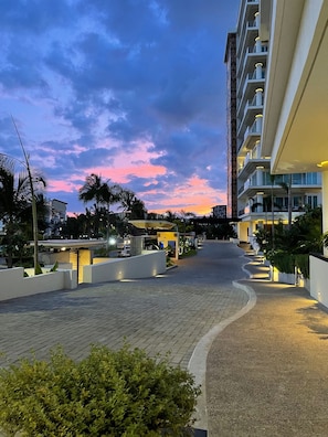 A hotel corridor with a curved pathway, palm trees, and a sunset sky.