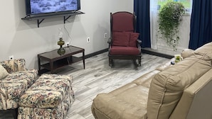 A living room with a television, a red armchair, a beige sofa, and a patterned ottoman. - Working farmette at converted warehouse in the Allegany Mountains. (Allegany)