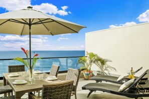 Un balcon avec une table dressée pour le dîner, des chaises en osier, un grand parasol et une vue sur l’océan.