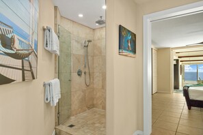 Une salle de bain avec une douche à parois de verre, un tableau représentant une scène de plage et une vue sur la mer par une porte ouverte.