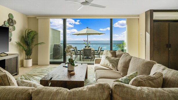 A living room with a sofa, a coffee table, and a ceiling fan. There is a large window with a view of the ocean. - Hawaii Life Rentals Presents Puu Poa 307 (Princeville)