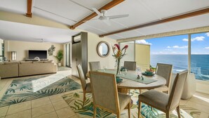 A spacious living room with a large TV, comfortable seating, a dining area with a table and chairs, and a view of the ocean. - Hawaii Life Rentals Presents Puu Poa 307 (Princeville)