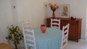 A dining area with a round table covered by a blue tablecloth, surrounded by four white chairs. A potted plant with yellow flowers is placed to the left of the table. - apartment in pretty village (Lumio)