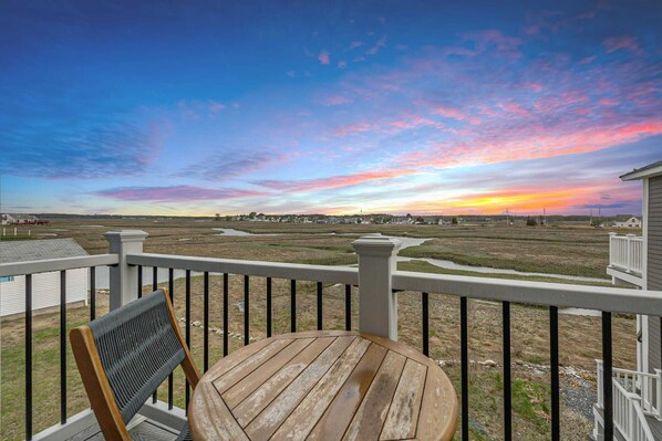 A balcony with a wooden table and chair, overlooking a flat landscape with distant buildings and a colorful sunset sky.