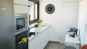 A compact kitchen with a Bosch refrigerator, microwave, and oven. There's a sink with a green towel, a clock, and a coffee machine on a small table. - Superb apartment with sea and mountain views (Canet-en-Roussillon)