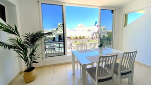 A room with a balcony, a dining table with chairs, a potted plant, and a view of a building with a blue roof. - Ocean Golf & Country Club A2 (Oasis del Sur)
