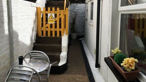 A narrow outdoor corridor with a yellow caution sign, a metal chair, a potted plant, and a staircase leading to a higher level. - Homely 2- bed Apartment -ground Floor-sleeps 4 (Ilfracombe)