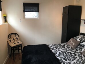 A bedroom with a bed, a chair, a wardrobe, a window with a blind, and a small table with a vase of flowers. - Homely 2- bed Apartment -ground Floor-sleeps 4 (Ilfracombe)