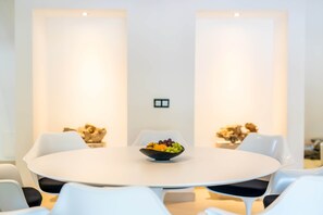 Uma sala de jantar moderna com uma mesa redonda, cadeiras brancas e uma cesta de frutas.