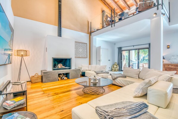A modern living room with a fireplace, a glass coffee table, and a high ceiling with exposed beams.