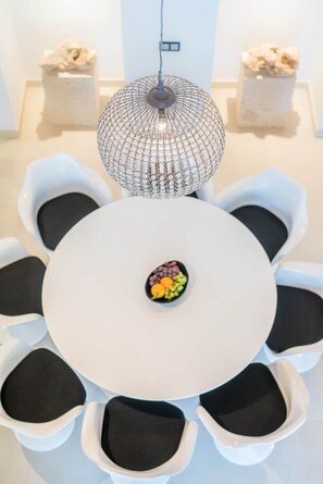 Uma mesa redonda com superfície branca e cadeiras estofadas em preto, uma luminária pendente decorativa e uma pequena tigela com frutas sobre a mesa.
