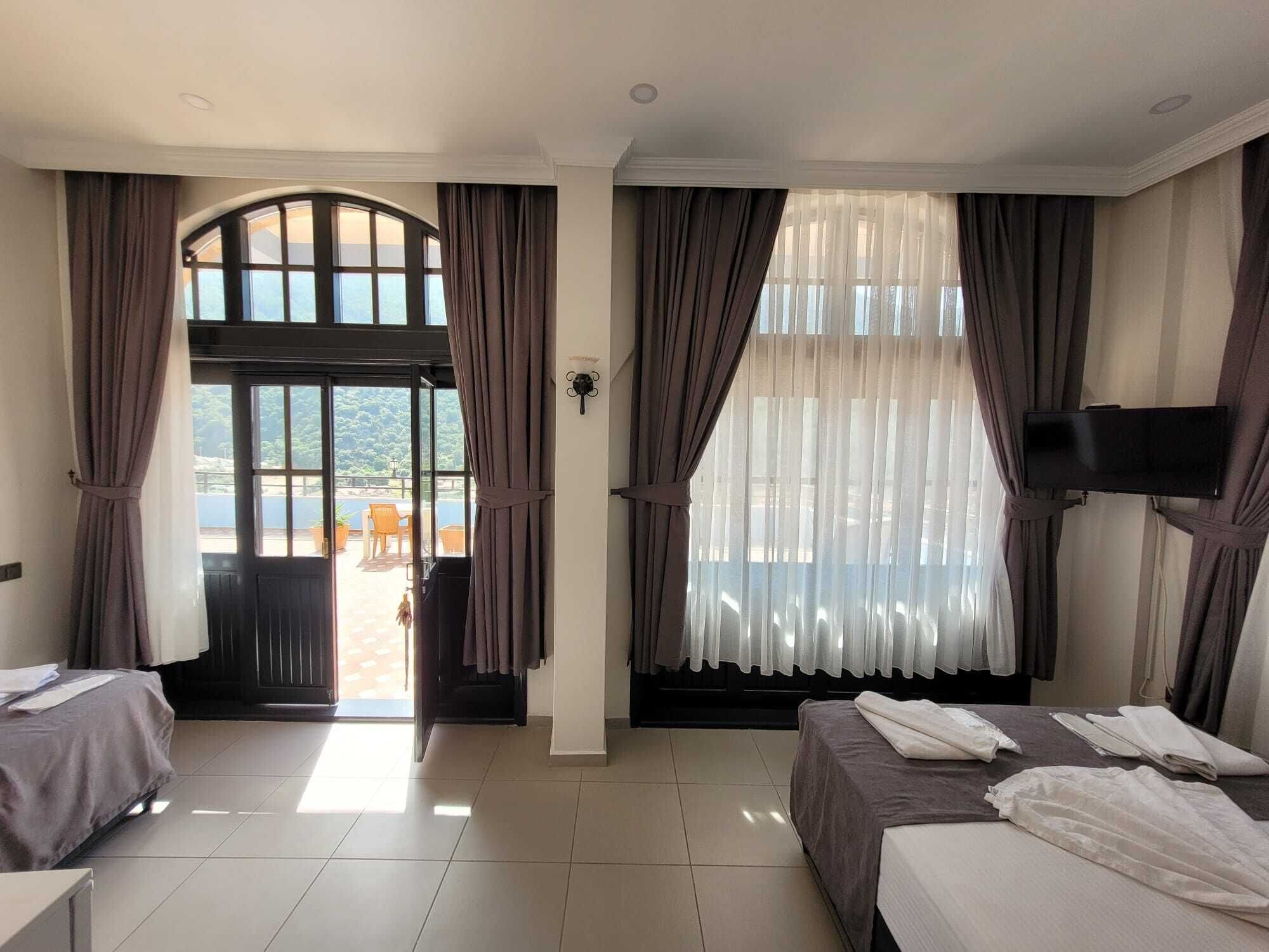 A hotel room with two beds, a TV mounted on the wall, a large window with curtains, and a view of the outdoors.