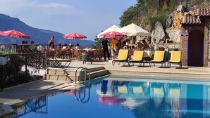 Ein Poolbereich mit Liegestühlen und Sonnenschirmen, ein Steinhaus mit Ziegeldach und eine gebirgige Landschaft im Hintergrund.