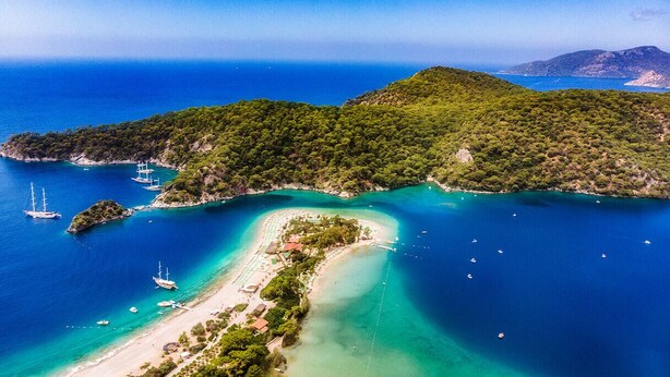 Luftaufnahme einer Küstenlandschaft mit einer kleinen Insel, klarem türkisblauem Wasser und einigen Booten.