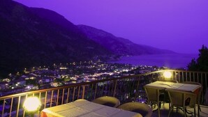 Ein Balkon mit Korbstühlen und einem Tisch, der auf eine Stadt und Berge in der Nacht blickt.