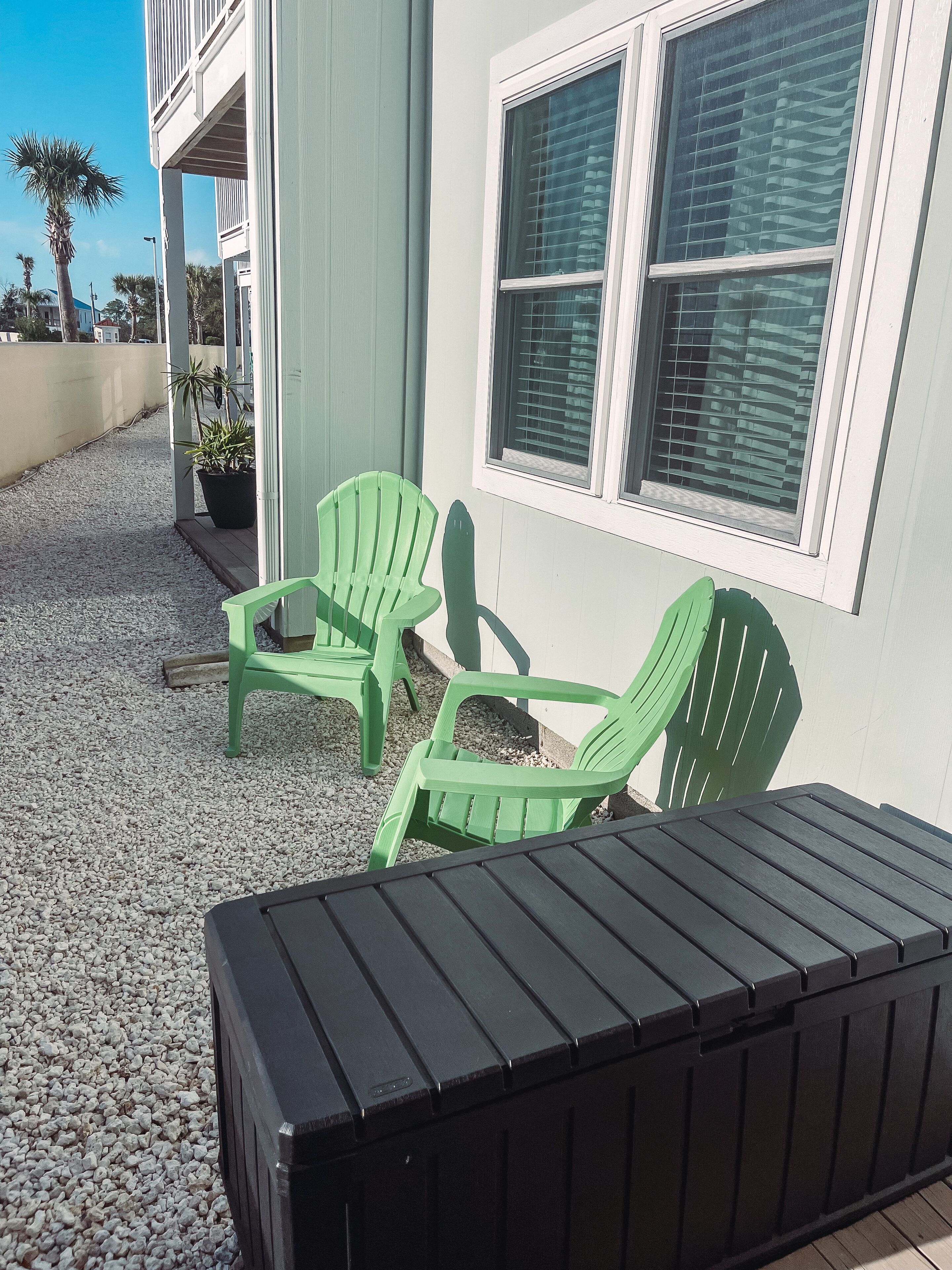 A patio with two green chairs and a black table, a building with a white exterior, and a window with blinds.