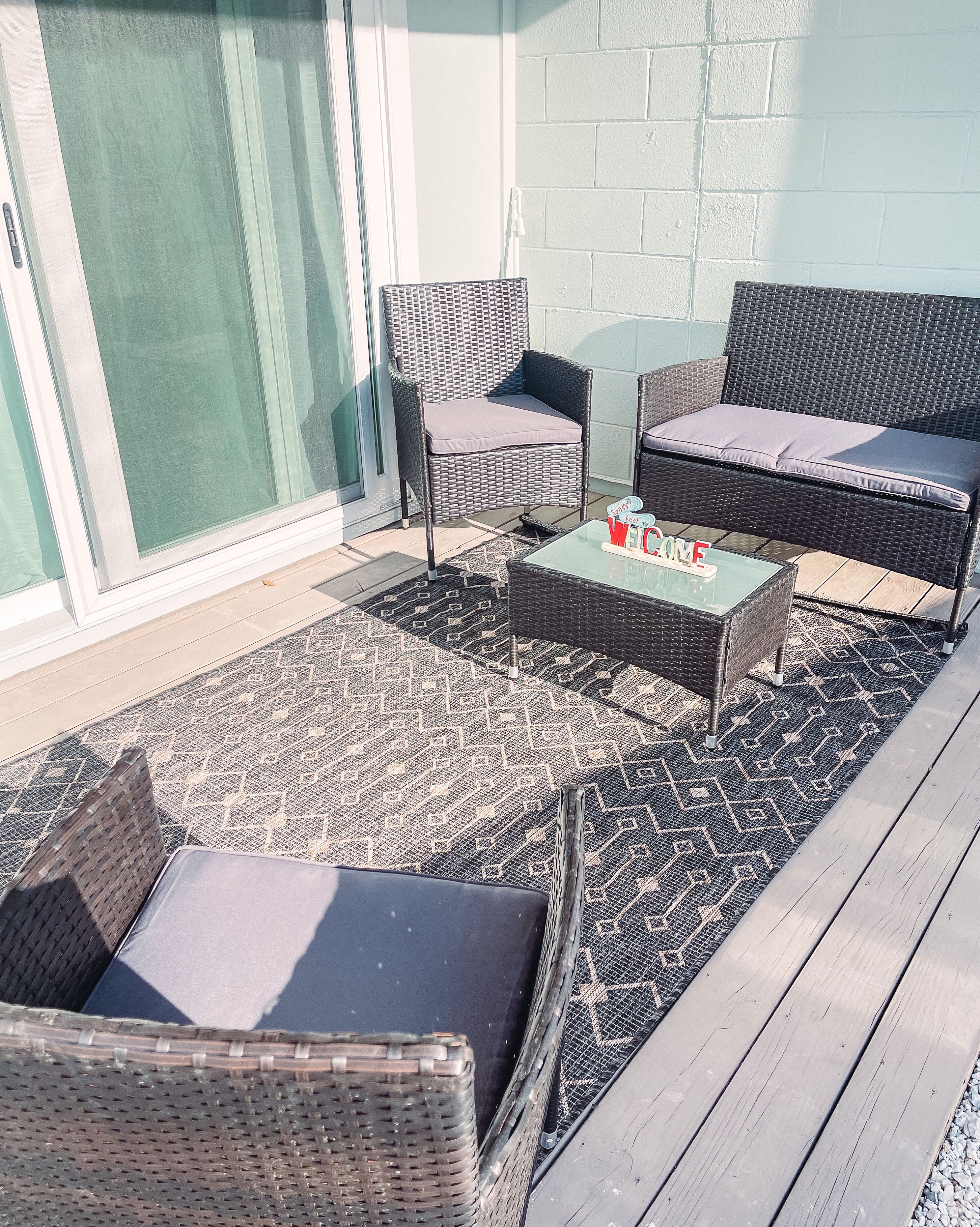 Una veranda esterna con mobili in vimini, un tavolino in vetro e un tappeto decorato.