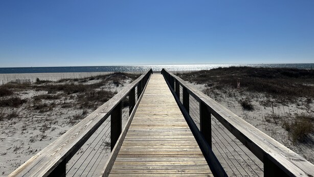 Une promenade en bois qui mène à une plage bordée de dunes de sable, sous un ciel bleu et dégagé.