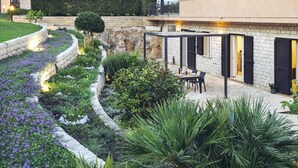 A modern outdoor patio area with a curved stone pathway, lush greenery, and a seating area with a table and chairs. - Isula Relax - Villa with pool (Modica)