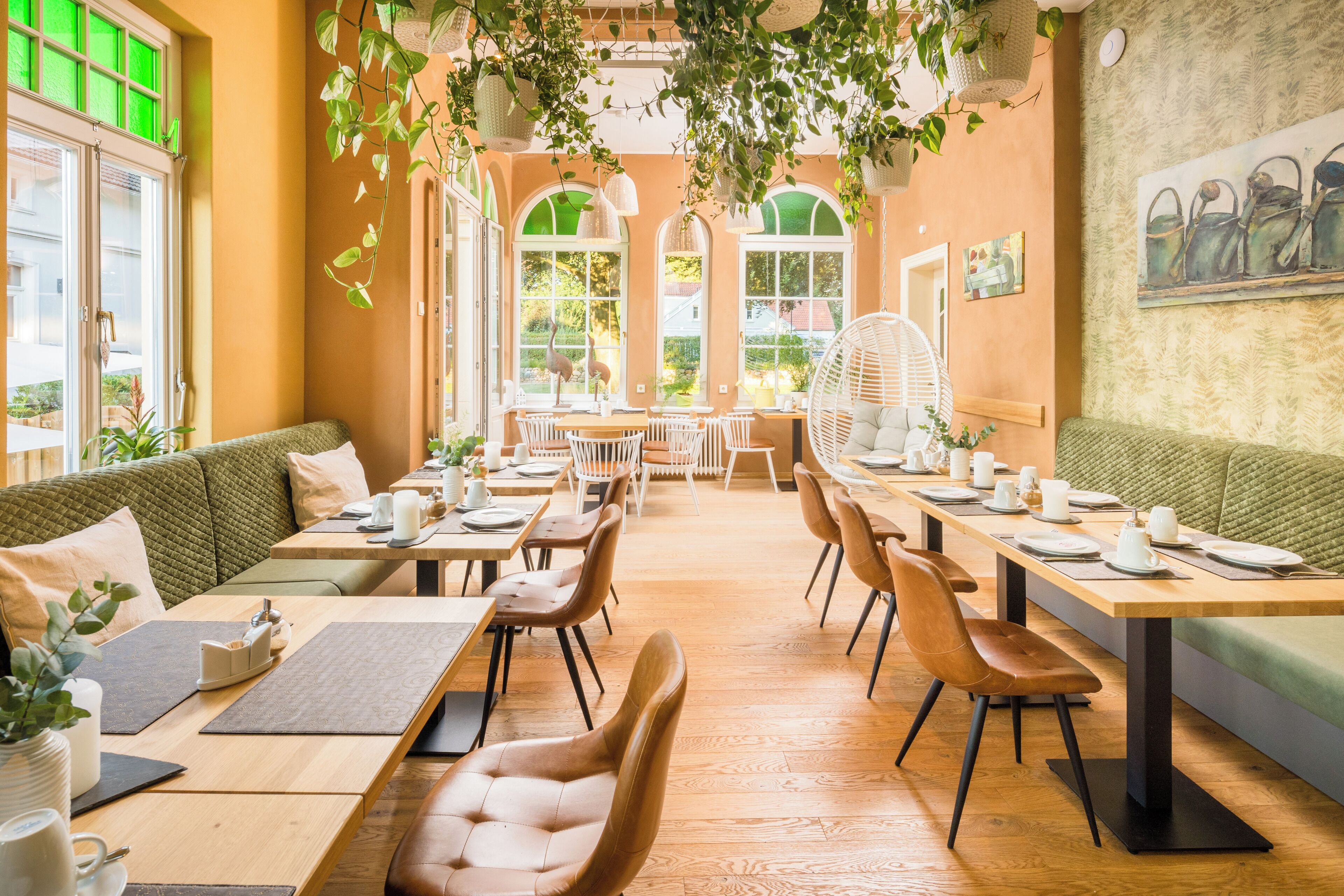 A cozy restaurant with wooden tables and chairs, green cushioned seating, and hanging plants.