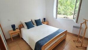 Une chambre avec un lit en bois, une literie blanche, deux oreillers bleus, un plaid bleu, une table de chevet avec une lampe et une fenêtre donnant sur un paysage verdoyant.